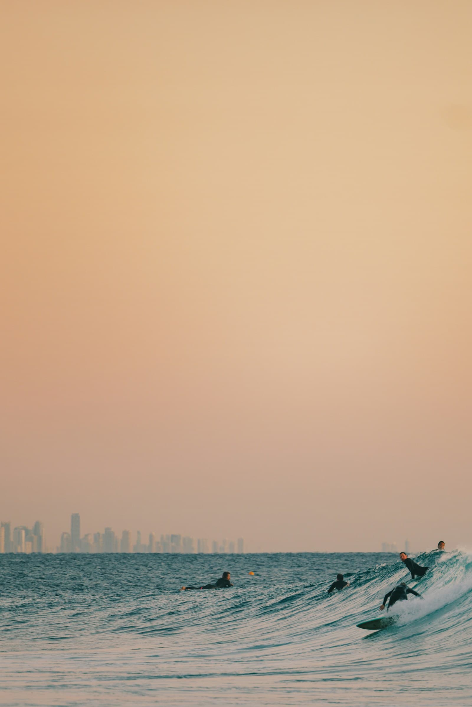 First-timer standing upright on a soft-top at Belongil, wide stance, small clean wave, instructor jogging alongside in the shallows. Late morning light, Cape Byron lighthouse just visible on the headland.