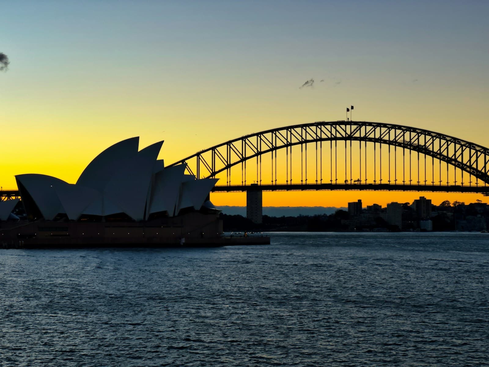Wide shot of the Sydney Harbour Bridge from Milsons Point at blue hour, Opera House visible bottom-right, ferry wake cutting through the frame, no people close to camera. Want weight and scale rather than the same postcard angle from Mrs Macquaries Point.